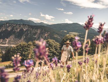 Eine Person in Latzhose geht auf einem Hügel mit lila Blumen und Bergen im Hintergrund.