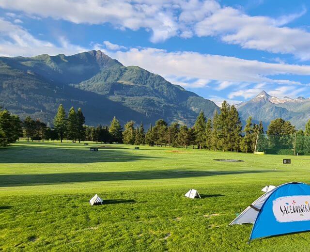 20210807_072100 Ein grüner Golfplatz mit einem blauen Zelt, Bäumen und Bergen unter einem blauen Himmel mit Wolken.