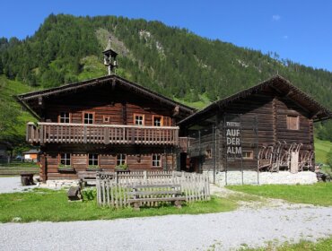 Zwei rustikale alpine Holzhäuser mit eingezäuntem Hof, umgeben von grünen Hügeln und blauem Himmel.
