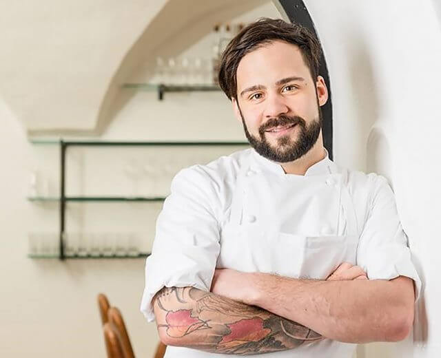 Ein lächelnder Chefkoch mit Bart und Tattoos steht mit verschränkten Armen in einer hellen, modernen Küche.
