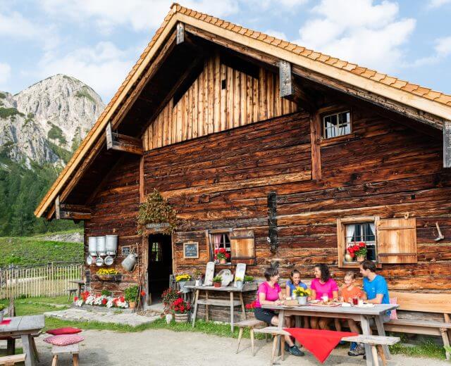 Eine Familie genießt eine Mahlzeit in einer Holzhütte mit Bergen und Bäumen im Hintergrund an einem sonnigen Tag.