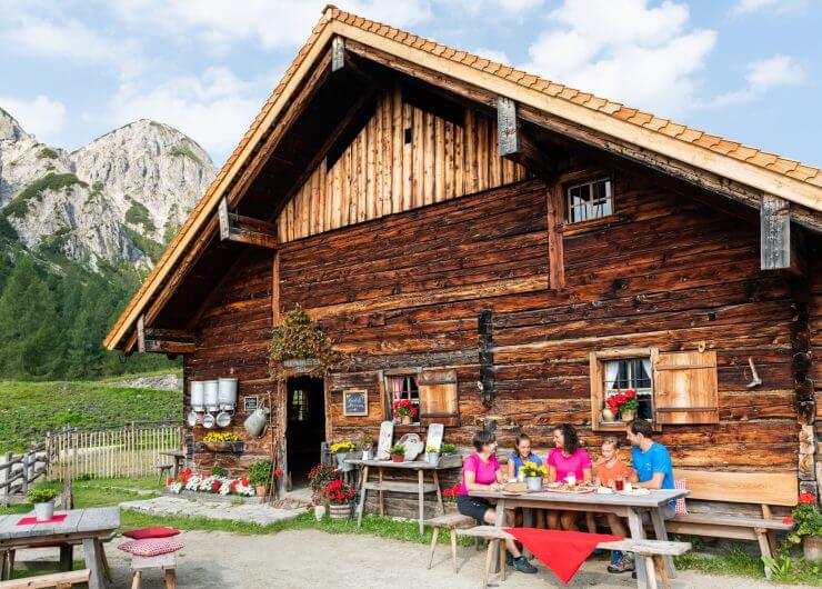 Eine Familie genießt eine Mahlzeit in einer Holzhütte mit Bergen und Bäumen im Hintergrund an einem sonnigen Tag.