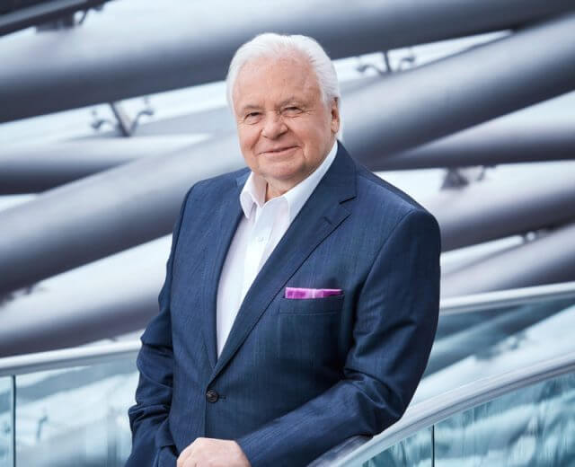 Ein älterer Mann im Anzug steht an einem Glasgeländer mit einem modernen architektonischen Hintergrund.