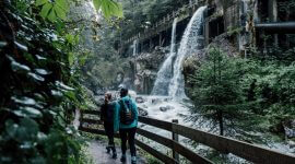 Seisenbergklamm © SalzburgerLand Tourismus Zwei Wanderer gehen auf einem Waldweg neben einem Fluss und einem Wasserfall, der aus einem alten Betonbauwerk fließt.