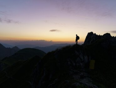 Ein Wanderer steht bei Sonnenuntergang auf einem Bergkamm und hebt sich von einem farbenfrohen Himmel ab.