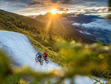 Zwei Radfahrer fahren bei Sonnenuntergang eine kurvenreiche Bergstraße hinauf, umgeben von Bäumen und malerischen Hügeln.