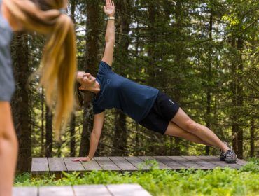Frau, die auf einer hölzernen Plattform in einem Wald einen Seitensprung macht; eine andere Person ist teilweise im Vordergrund zu sehen.
