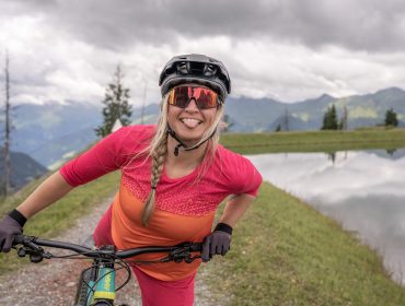 Eine lächelnde Frau mit Helm und Sonnenbrille posiert spielerisch mit ihrem Fahrrad an einem Bergsee.