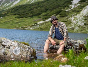 Ein Mann sitzt an einem See, die Füße im Wasser, umgeben von Felsen und grünen Hügeln.