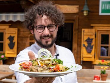 Ein Mann mit lockigem Haar und Brille präsentiert einen Teller mit Essen im Freien vor einem rustikalen Holzgebäude.