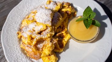 Ein Teller Kaiserschmarrn mit Puderzucker, serviert mit Apfelmus und einer Minzgarnitur auf einem rustikalen Holztisch.