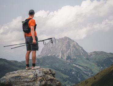 DSC09233 Ein Wanderer in Orange steht auf einem Felsen und blickt auf einen entfernten Berg unter einem bewölkten Himmel.