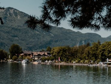 Ein ruhiger See mit Bäumen, Häusern und Bergen im Hintergrund unter einem klaren Himmel.