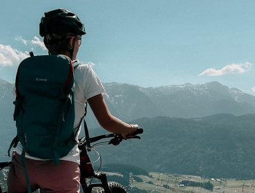 Person mit Helm und Rucksack auf einem Mountainbike, mit Blick auf eine malerische Berglandschaft unter klarem Himmel.