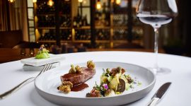 Elegantes Gourmet-Menü mit Rotwein auf einem weißen Tischtuch in einem schummrigen, gehobenen Restaurant.