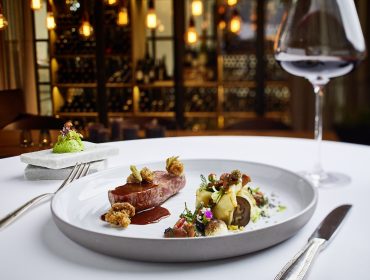 Elegantes Gourmet-Menü mit Rotwein auf einem weißen Tischtuch in einem schummrigen, gehobenen Restaurant.