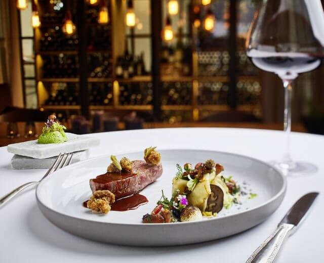 Rauriser Reh von Vitus Cooking Elegantes Gourmet-Menü mit Rotwein auf einem weißen Tischtuch in einem schummrigen, gehobenen Restaurant.