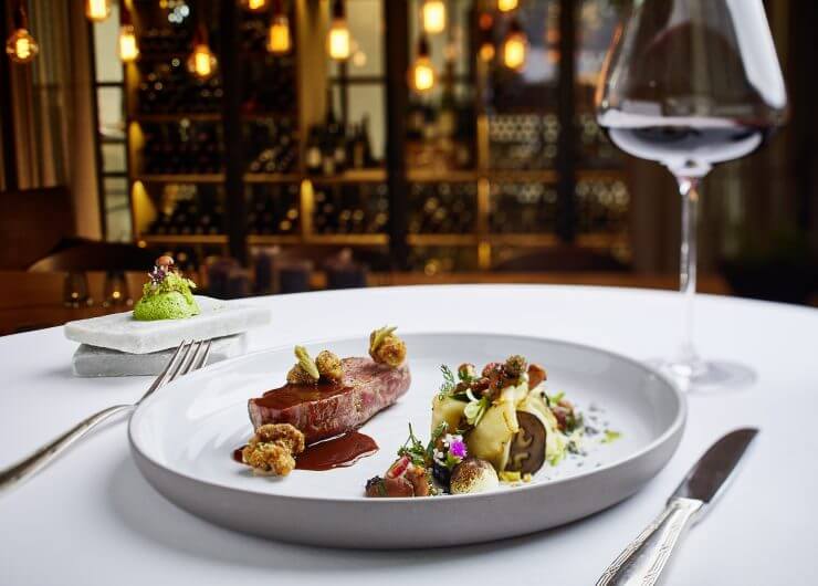 Elegantes Gourmet-Menü mit Rotwein auf einem weißen Tischtuch in einem schummrigen, gehobenen Restaurant.