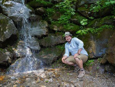 Ein Mann in Shorts und mit Hut hockt an einem kleinen Wasserfall, der von Felsen und Grün umgeben ist.