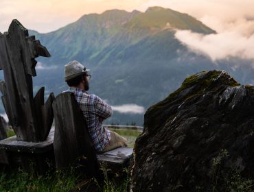 Eine Person mit Hut sitzt auf einer rustikalen Holzbank und blickt auf die Berge unter einem bewölkten Himmel.