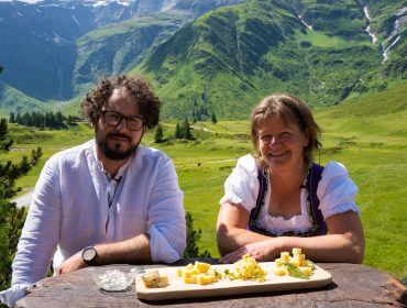 Zwei Personen im Freien an einem Holztisch mit Käse; Berge und grüne Landschaft im Hintergrund.