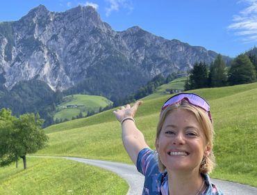 Eine lächelnde Frau zeigt auf eine Bergkette an einem sonnigen Tag in einem üppig grünen Tal.