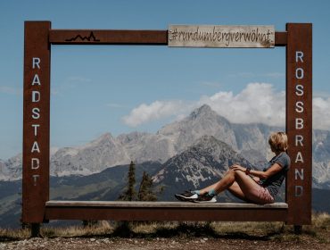 Eine Frau sitzt auf einer Bank innerhalb eines großen Metallrahmens mit Bergen und blauem Himmel im Hintergrund.
