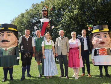 Im Freien posierende Gruppe in traditioneller österreichischer Tracht mit zwei großen Maskottchen und einer großen Soldatenfigur.