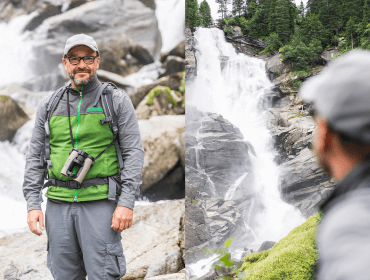 Ein Mann in Outdoor-Ausrüstung steht an einem Felsen und blickt auf einen großen Wasserfall und einen Wald im Hintergrund.