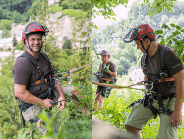 Zwei Männer mit Helmen und Klettergurten seilen sich an einem üppigen, grünen Hang mit einer Stadt im Hintergrund ab.