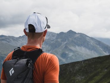 Eine Person in orangefarbenem Hemd und weißer Mütze blickt auf Berggipfel unter einem bewölkten Himmel.