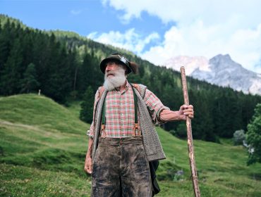 Älterer Mann mit Bart und Spazierstock steht in einer grasbewachsenen Berglandschaft mit Bäumen und Gipfeln im Hintergrund.