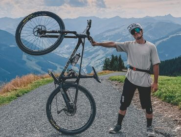 Mann in Radfahrerkleidung, der ein Mountainbike an einem Rad auf einem malerischen Bergpfad aufrecht hält.