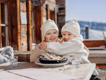Zwei lächelnde Kinder in Winterkleidung umarmen sich an einem Tisch im Freien mit einem Dessert und verschneiten Bergen im Hintergrund.
