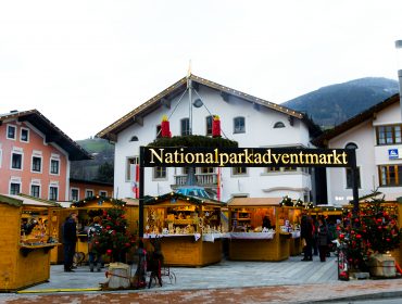 Hölzerne Weihnachtsmarktstände vor alpinen Gebäuden mit der Aufschrift Nationalparkadventmarkt.