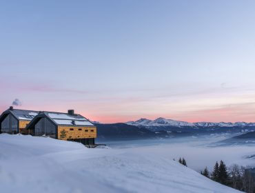 Ein modernes Haus in verschneiten Bergen bei Sonnenaufgang, mit nebligen Tälern und rosa Himmel im Hintergrund.