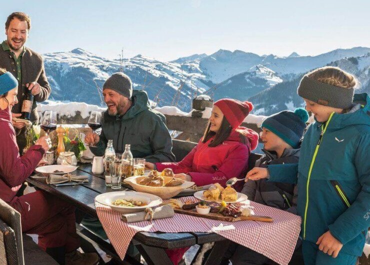 Fünf Personen in Winterkleidung genießen eine gemeinsame Mahlzeit an einem Tisch im Freien mit verschneiten Bergen im Hintergrund.