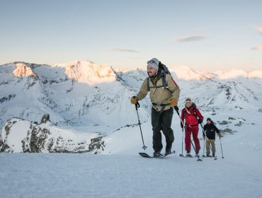 Drei Personen in Winterkleidung wandern mit Schneeschuhen in einer verschneiten Berglandschaft bei Sonnenaufgang oder Sonnenuntergang bergauf.