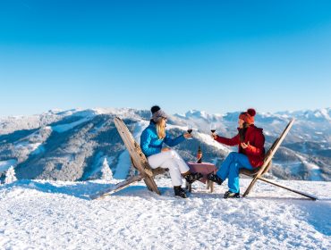 Zwei Menschen stoßen mit Gläsern an, während sie in Stühlen auf einem verschneiten Berg mit malerischen Gipfeln im Hintergrund sitzen.