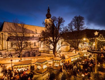 Festlicher Weihnachtsmarkt mit Lichtern, Holzbuden und einer großen Kirche in der Abenddämmerung, umgeben von Winterbäumen.