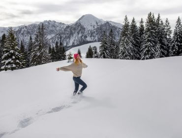 Eine Person mit einem rosa Hut läuft fröhlich durch den tiefen Schnee und setzt sich für Nachhaltigkeit inmitten von Bergen und Kiefern ein.