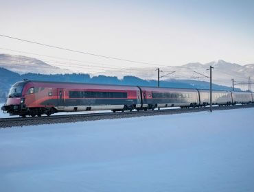 Ein rot-weißer Personenzug ÖBB fährt durch eine verschneite Landschaft mit Bergen im Hintergrund.