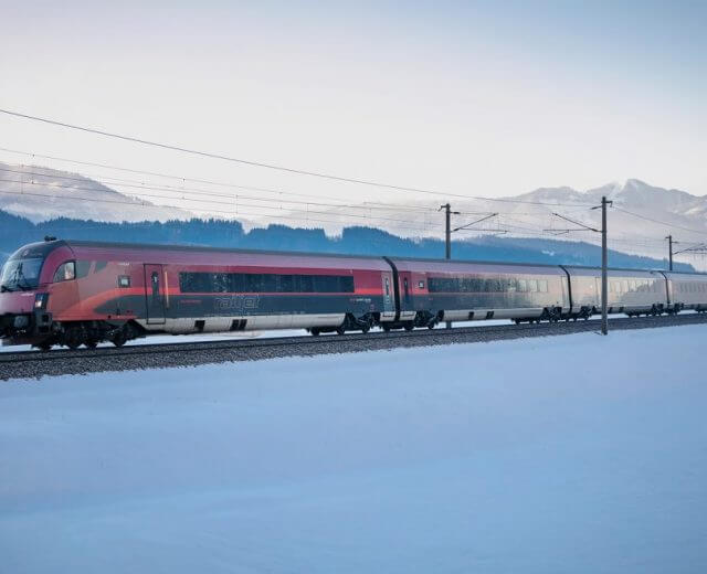 Bahn im Winter (c) OEBB Ein rot-weißer Personenzug ÖBB fährt durch eine verschneite Landschaft mit Bergen im Hintergrund.
