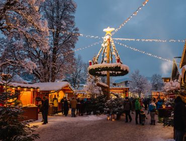 In der Abenddämmerung genießen die Menschen in Altenmarkt-Zauchensee einen festlichen Weihnachtsmarkt unter freiem Himmel, mit verschneiten Bäumen und Lichterketten.