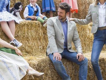 Fünf Erwachsene und Kinder in traditioneller österreichischer Kleidung sitzen und stehen auf Heuballen im Freien und lächeln.