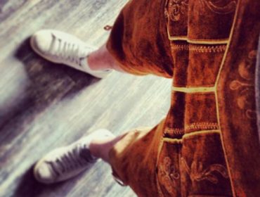 Person in braunen Lederhosen und weißen Turnschuhen, die auf einem Holzboden steht, von oben gesehen.