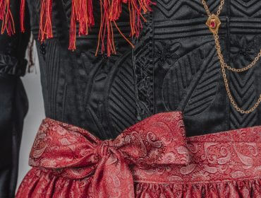 Nahaufnahme eines traditionellen schwarzen Kleides mit roten Fransen, einer rot gemusterten Schärpe und mehrlagigen goldenen Halsketten.
