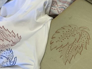 Weißes T-Shirt mit Blattmotiven neben Skizzenpapier mit ähnlichen Blattmustern und bunten Textilien.