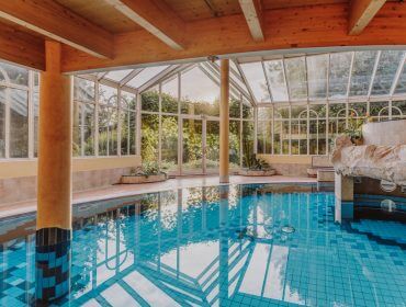 Hallenbad im Gschirnwirt mit blauen Fliesen, Glaswänden, Holzbalken und Sonnenlicht, das durch die Fenster fällt.