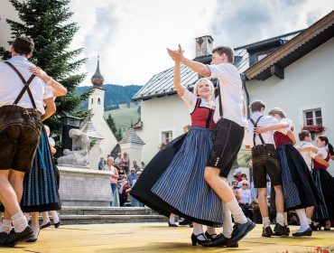 Menschen in traditioneller österreichischer Kleidung tanzen im Freien auf einer Holzbühne vor den Augen des Publikums.